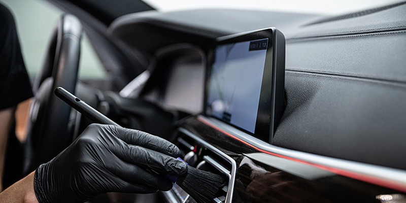 Gloved hand using a detailing brush to clean the air vent and dashboard area of a car interior, with a digital display screen in view.